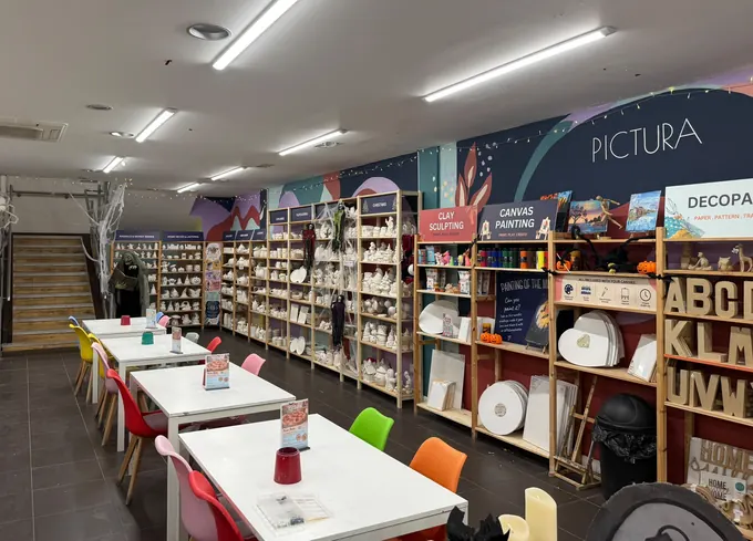Inside Pictura Chester: white painting tables with colourful chairs, shelves of pottery ready to paint and the PICTURA wall mural